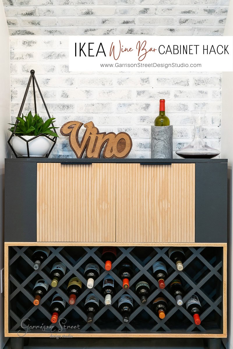 Stylish wine bar showcasing an IKEA wine bar cabinet hack with a black and wood design. Wine bottles are arranged in a cross-grid shelf, complemented by a small plant in a geometric holder, a wooden "Vino" sign, and two bottles topped with a glass.