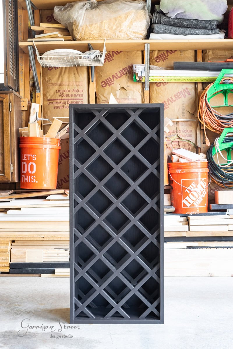 A black wooden wine rack with a lattice design stands upright in a garage or workshop, reminiscent of an IKEA Wine Bar Cabinet hack. Surrounding the rack are various tools, wooden planks, and orange buckets. Shelves with materials are visible in the background.