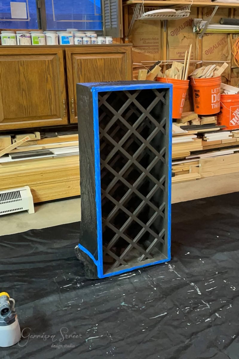 In the workshop, an IKEA wine rack undergoes a stylish transformation into a chic wine bar cabinet. Painted black with precision, its crisscross design is neatly outlined by blue tape. Tools and paint surround this innovative hack, promising a sleek addition to any decor.