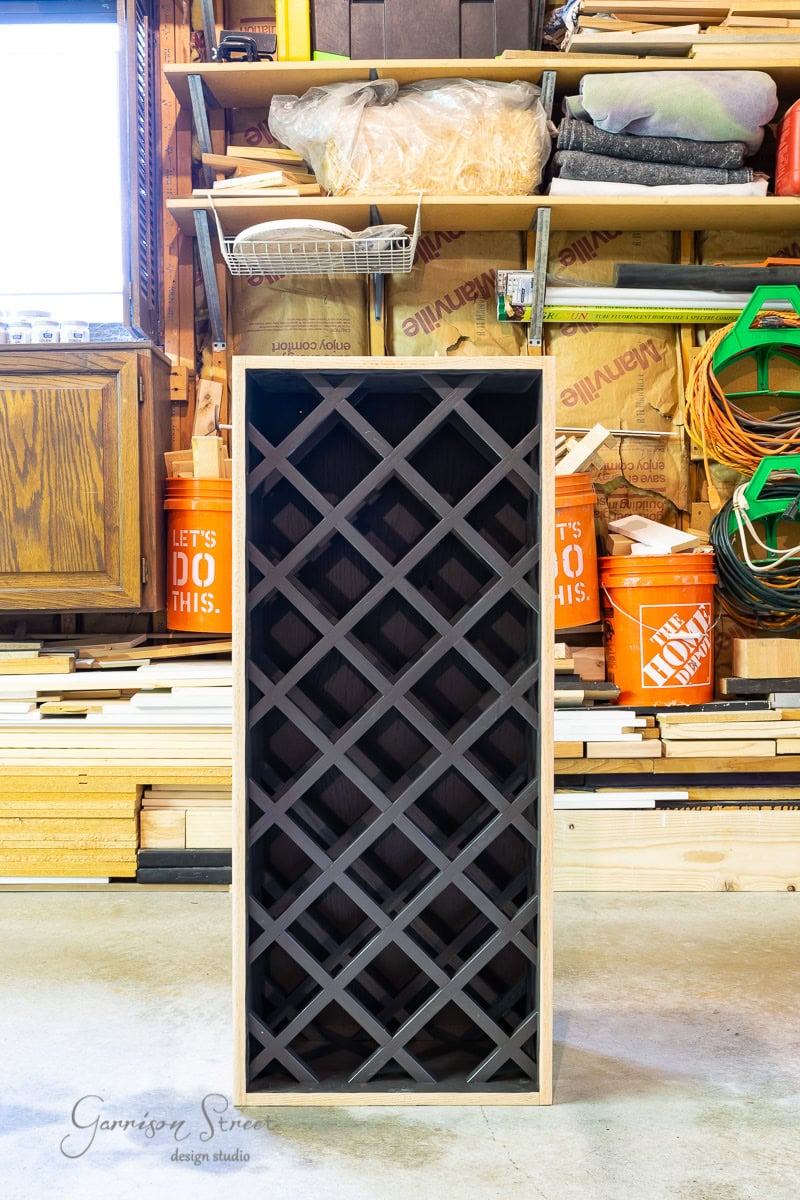 A wooden wine rack with a black grid design stands upright in a cluttered garage, resembling an IKEA Wine Bar Cabinet hack. The background features Home Depot buckets, stacked wood, tools, and shelves.