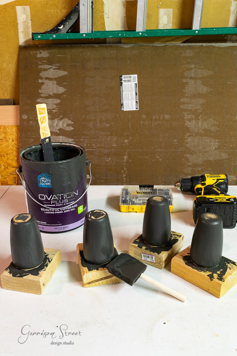 Tools for a DIY project are on display: a paint can with a stirring stick, three black-painted cups upside down on wood blocks, sponge brushes, and a power drill with screws. A protective sheet covers the background—a perfect setup for your next IKEA wine bar cabinet hack.
