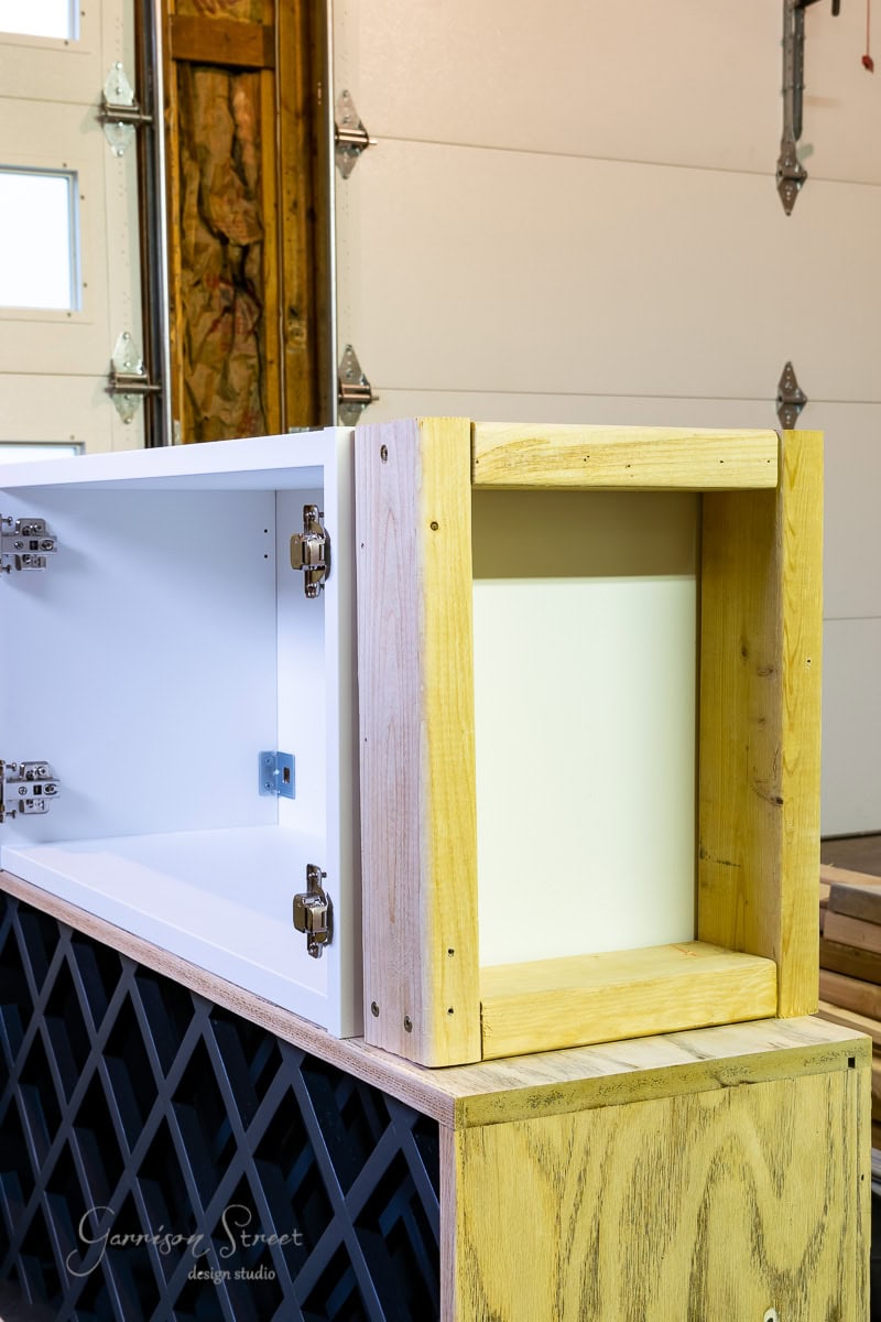 In the workshop, a wooden cabinet frame is under construction, showcasing lattice detailing and exposed hinges. This IKEA hack combines sections of white and natural wood, reimagining it as an elegant wine bar. Visible in the background are the garage door and various tools.