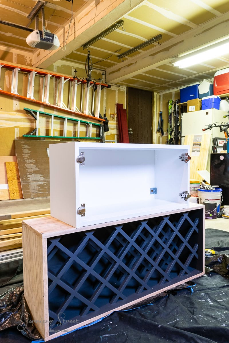 A garage workshop showcases a DIY IKEA wine bar cabinet hack, with a completed wooden cabinet featuring a black lattice pattern on the bottom and open white storage space on top. Various tools, ladders, and materials are scattered in the background, hinting at ongoing creative projects.