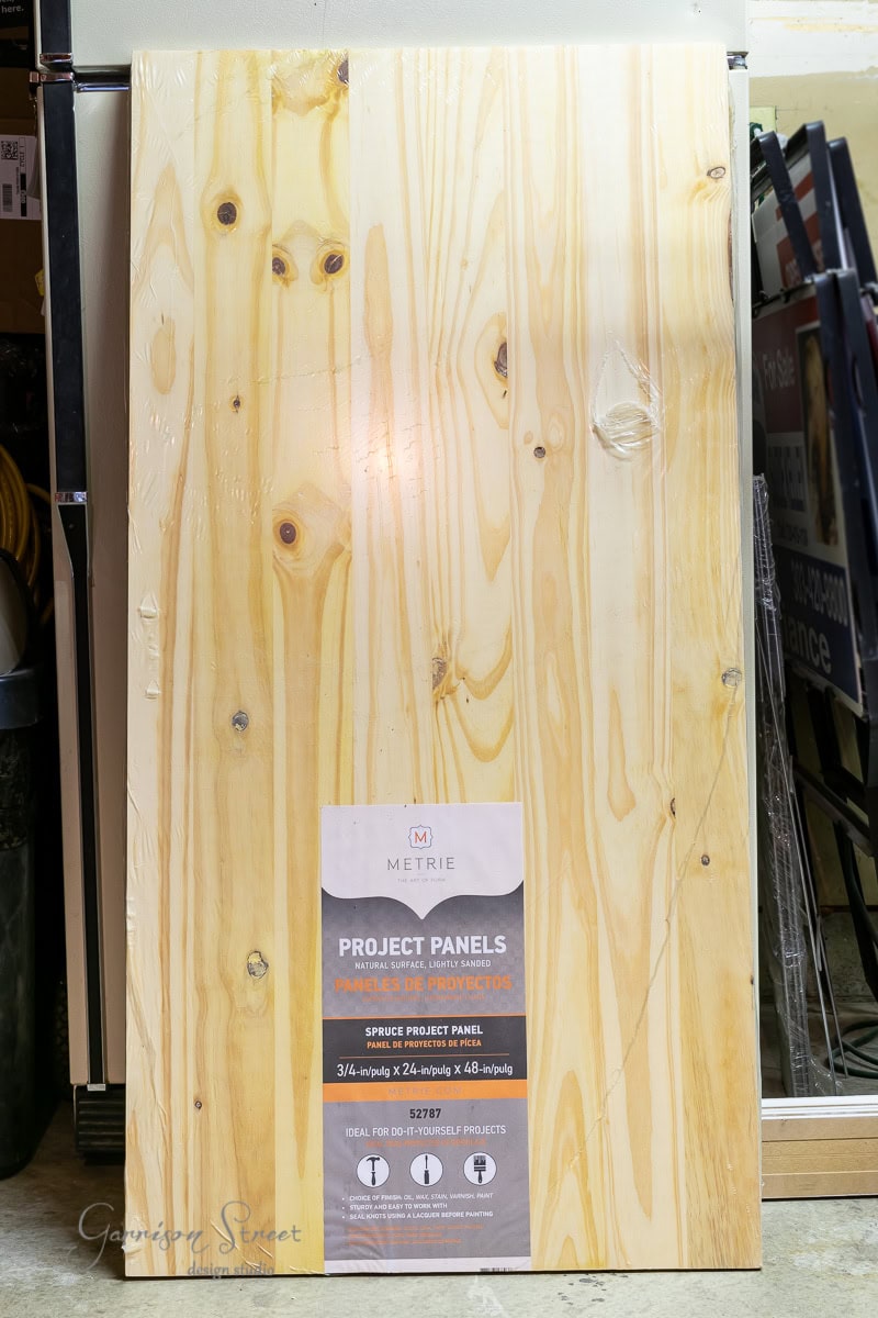 A large, rectangular spruce project panel with visible wood grain stands upright, wrapped in clear plastic. Labeled by "Metrié" for its dimensions and DIY potential, it's a perfect start for your next IKEA Wine Bar Cabinet hack.