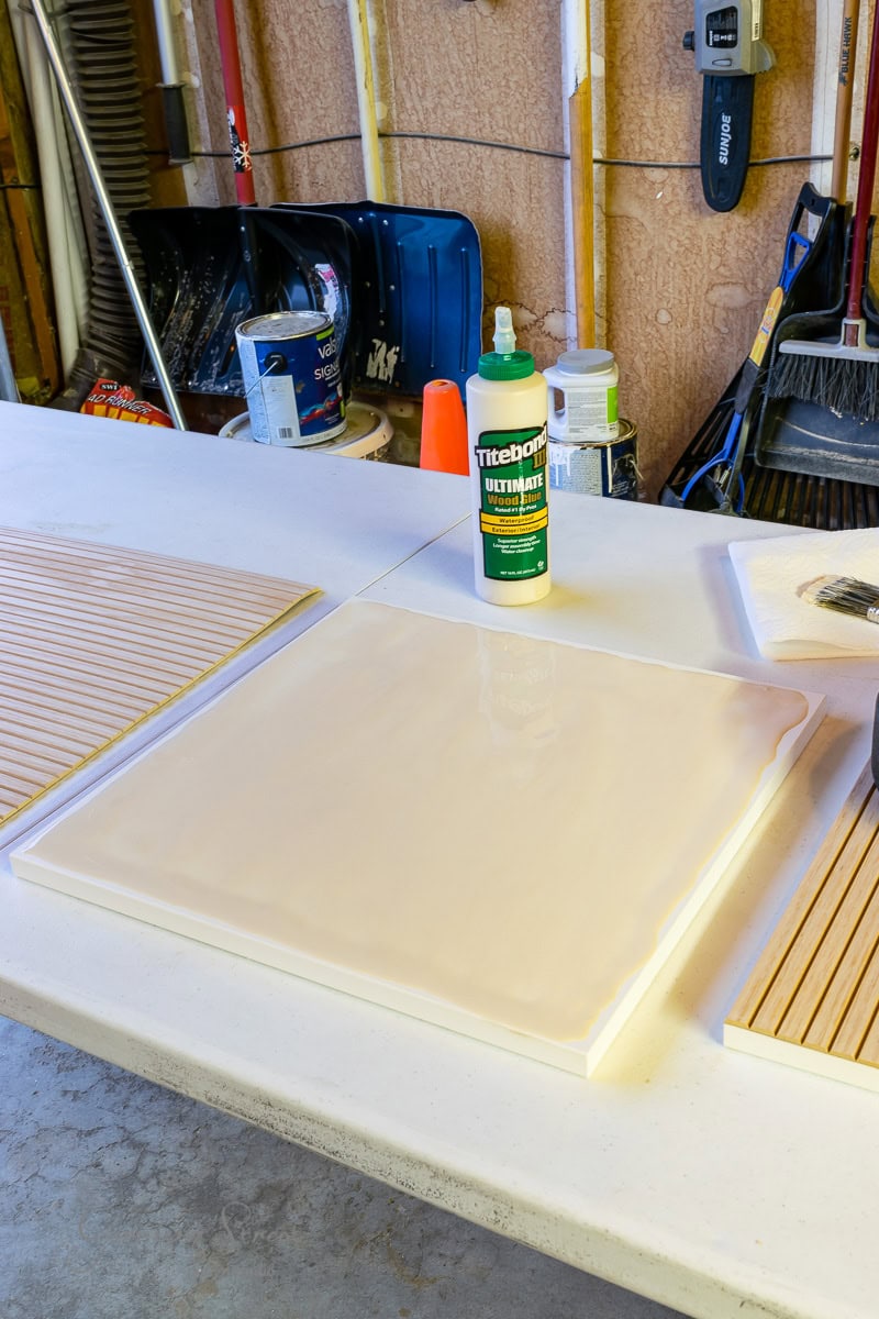 A workshop table with a wooden panel coated in glue showcases an IKEA Wine Bar Cabinet Hack in progress. The Titebond Ultimate Wood Glue stands nearby, surrounded by various tools and materials like paint cans and brooms, hinting at the creativity unfolding.