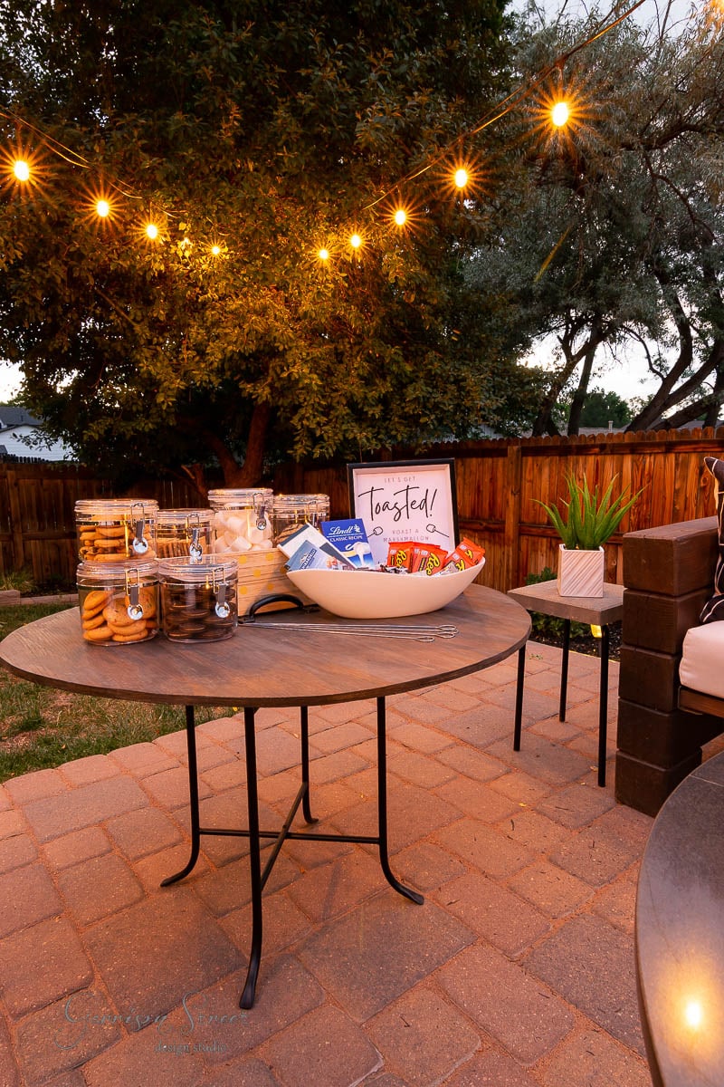 A cozy outdoor patio with string lights illuminating a round table featuring a backyard s'mores bar, complete with a bowl of snacks, jars of cookies, and a framed sign reading "Toasted!" A potted plant decorates the corner, set against trees and a wooden fence.
