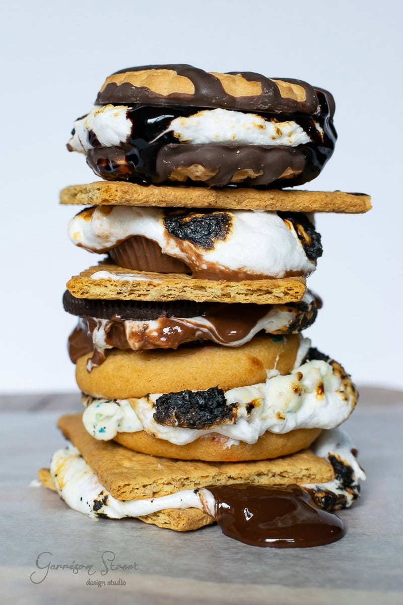 A visually appealing stack of s'mores from a backyard s'mores bar features toasted marshmallow, melted chocolate, and peanut butter cups sandwiched between graham crackers and cookies. Some chocolate is deliciously dripping down the sides.