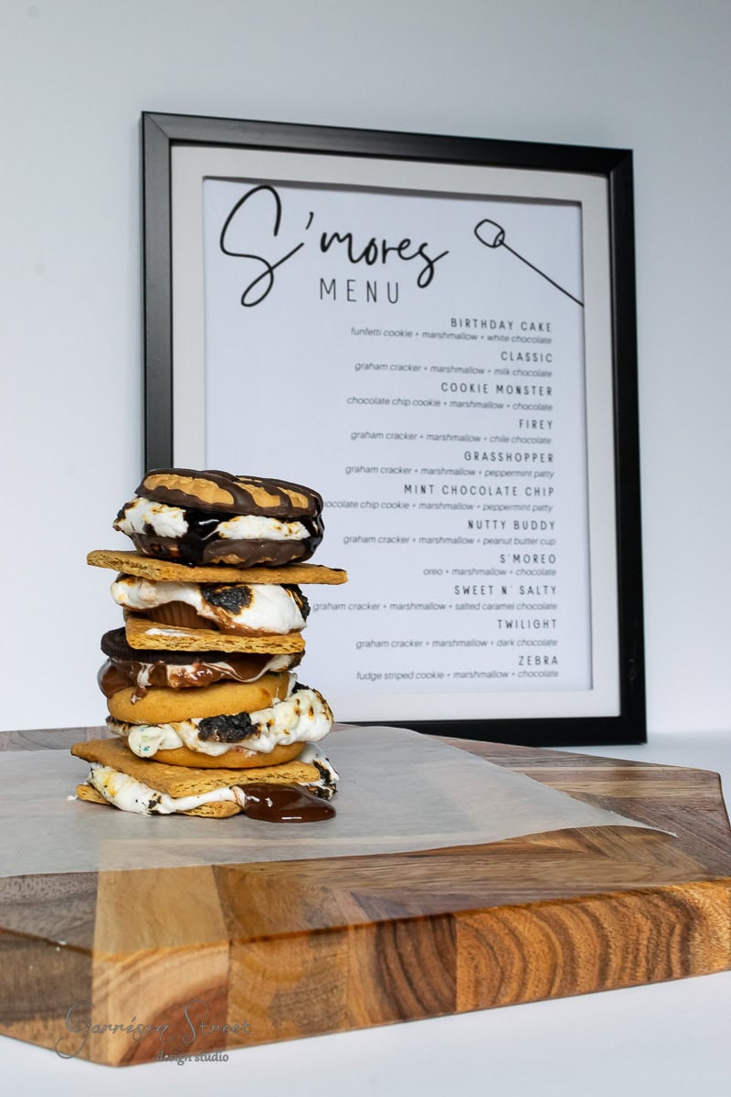 A stack of s'mores with different fillings sits enticingly on a wooden board, perfectly capturing the vibe of a backyard s'mores bar. Behind them, a framed "S'mores Menu" lists exciting flavors like Birthday Cake and Cookie Monster.