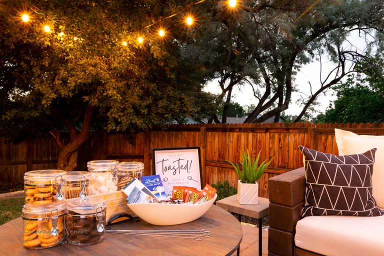 A cozy backyard s'mores bar setup with string lights overhead. A wooden table holds a bowl of chocolate, marshmallows, and graham crackers alongside glass jars. A cushioned couch is nearby, while trees gently border the wooden fence.