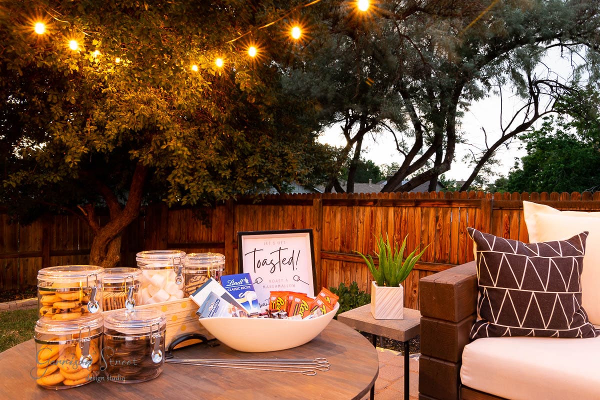 A cozy backyard s'mores bar setup with string lights overhead. A wooden table holds a bowl of chocolate, marshmallows, and graham crackers alongside glass jars. A cushioned couch is nearby, while trees gently border the wooden fence.