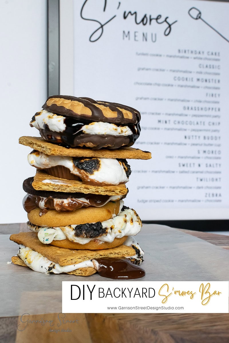 A stack of creative s'mores with various toppings, including chocolate and marshmallow, is displayed on a wooden surface. The enticing s'mores menu entices you to create your own masterpiece at this DIY backyard s'mores bar, as the words "DIY Backyard S'mores Bar" beckon from below.
