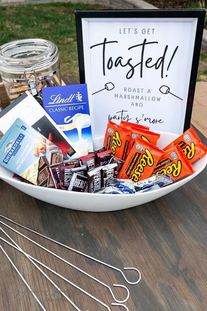 A backyard s'mores bar with a sign declaring "Let's Get Toasted! Roast a Marshmallow and Party S'more" offers a charming setup. The tray brims with Reese's, Hershey's, and graham crackers, alongside skewers and a jar of fluffy marshmallows, inviting endless delicious creations under the open sky.