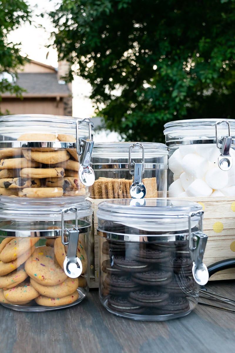 Clear jars filled with cookies, graham crackers, and marshmallows are arranged on a wooden surface, creating the perfect backyard s'mores bar. The background features green foliage, adding a natural touch to the outdoor setting.