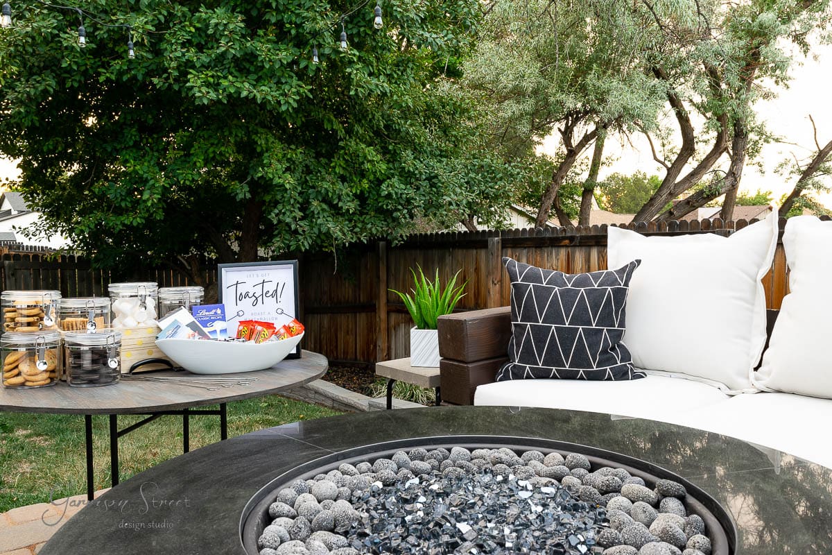 A cozy outdoor seating area features a cushioned sofa with patterned pillows, a fire pit perfect for a backyard s'mores bar in the foreground, and a side table with jars of snacks and a bowl of chips labeled "Tasty!" under string lights and trees.