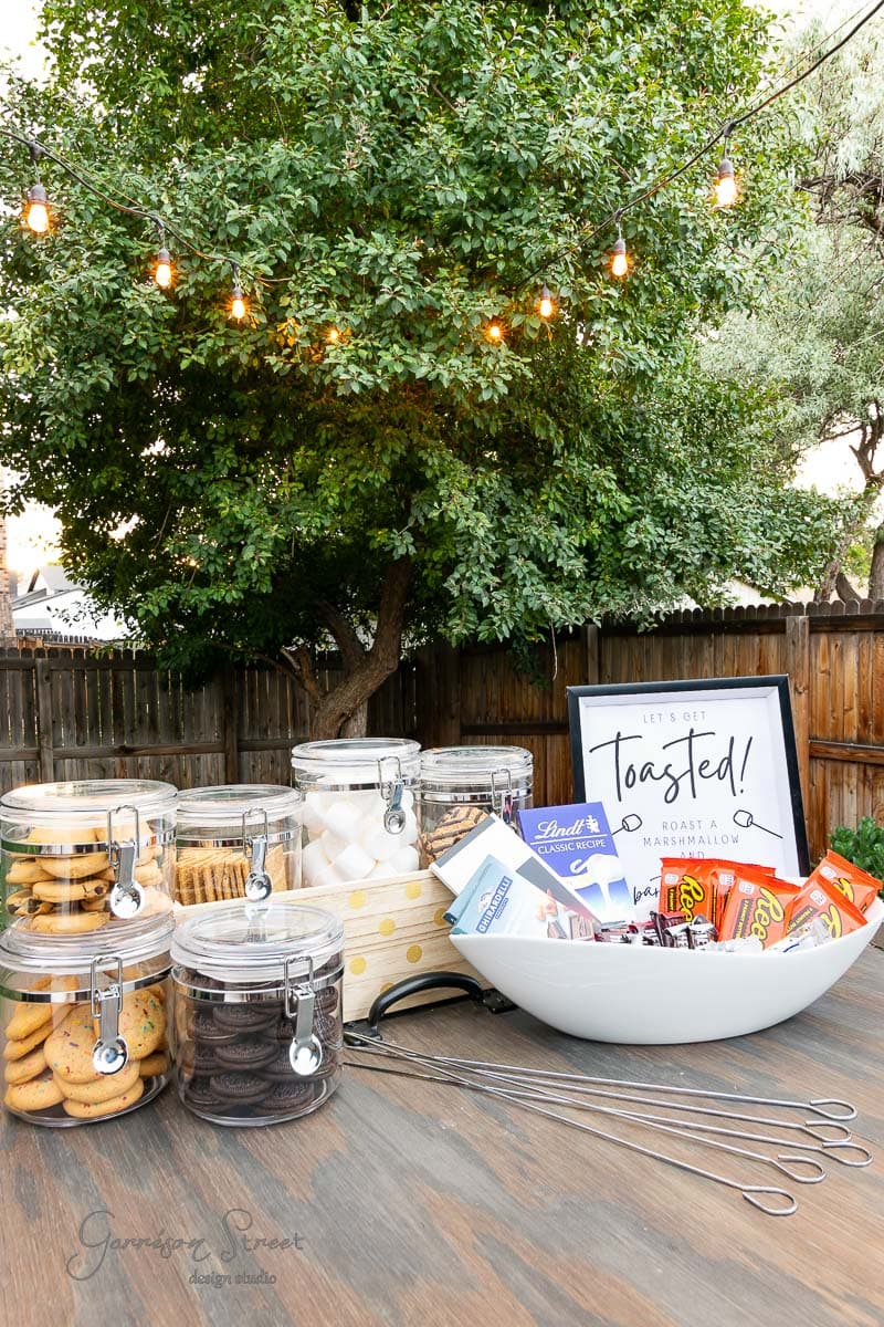 An outdoor setup boasts a wooden table transformed into a backyard s'mores bar. Jars of cookies, marshmallows, and chocolates tempt guests. A large bowl holds more treats beneath a "Let's Get Toasted" sign. String lights twinkle above, with a tree and wooden fence setting the perfect scene.
