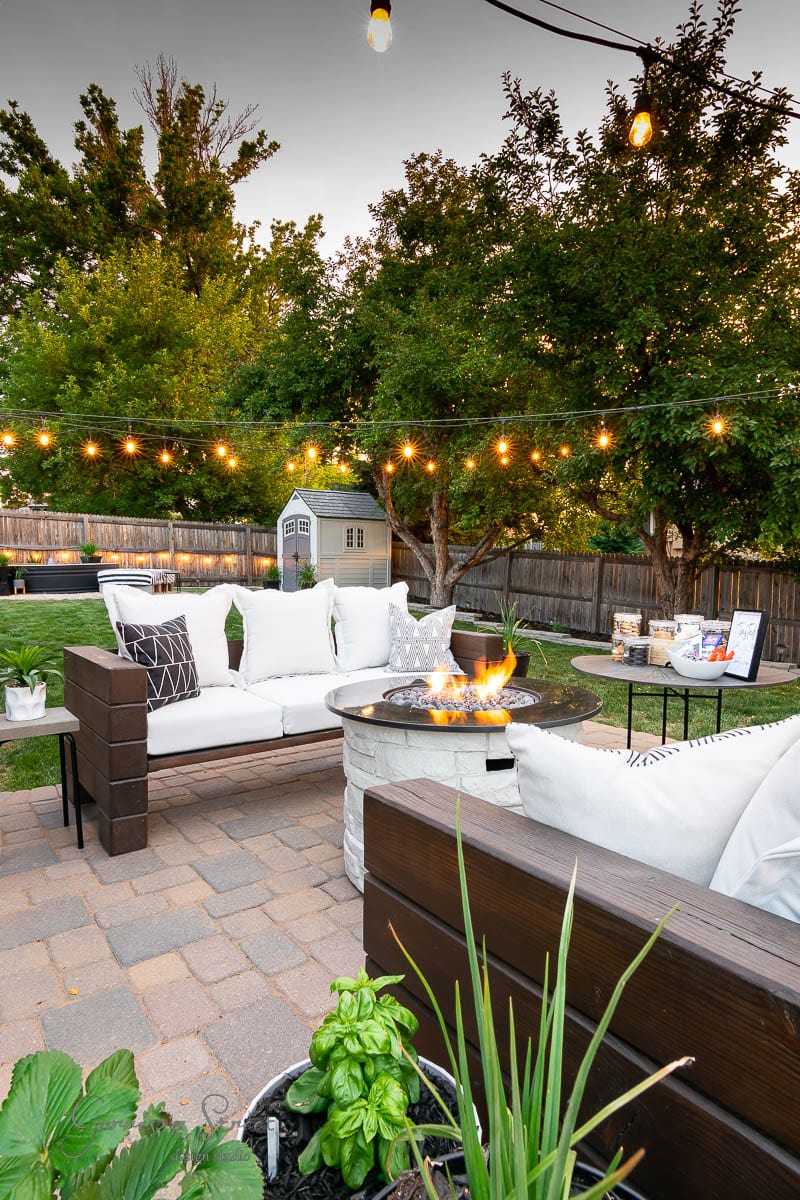 A cozy outdoor patio with white cushioned seating around a fire pit, perfect for a backyard s'mores bar. String lights illuminate the area, while lush greenery and a small playhouse complete the scene. Potted plants are charmingly placed in the foreground.