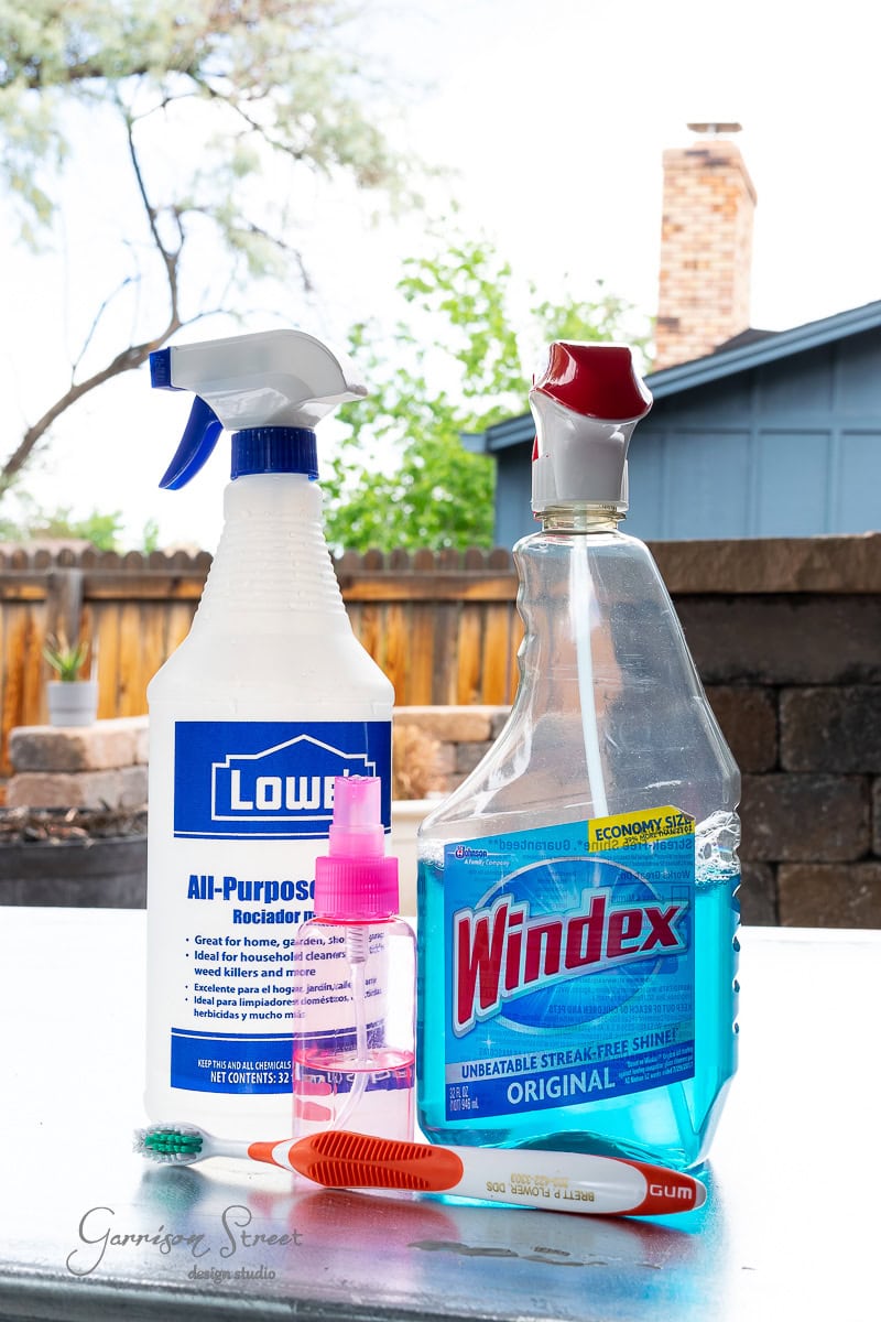 A group of cleaning supplies, including a spray bottle of all-purpose cleaner, a bottle of Windex, a small pink spray bottle, and a toothbrush, is arranged on a stock tank outdoors. A wooden fence and trees grace the garden backdrop.