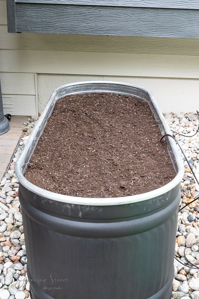 An outdoor metal planter resembling a stock tank is filled with fresh soil, placed on a gravel area beside a building. The rectangular garden planter appears ready for planting.