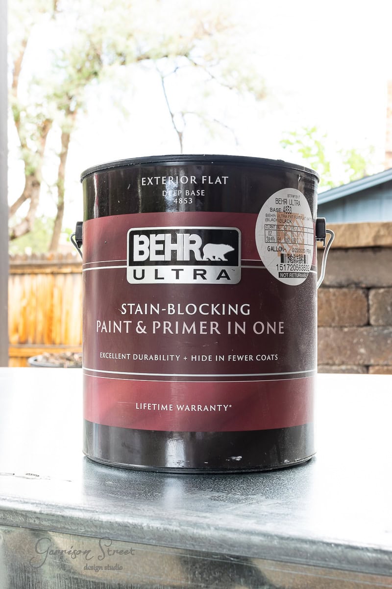A large can of Behr Ultra stain-blocking paint and primer in one rests on a metal stock tank outdoors. The can is labeled "Exterior Flat: Ultra Pure White," boasting excellent durability and a lifetime warranty. A wooden fence and lush garden provide the perfect backdrop.
