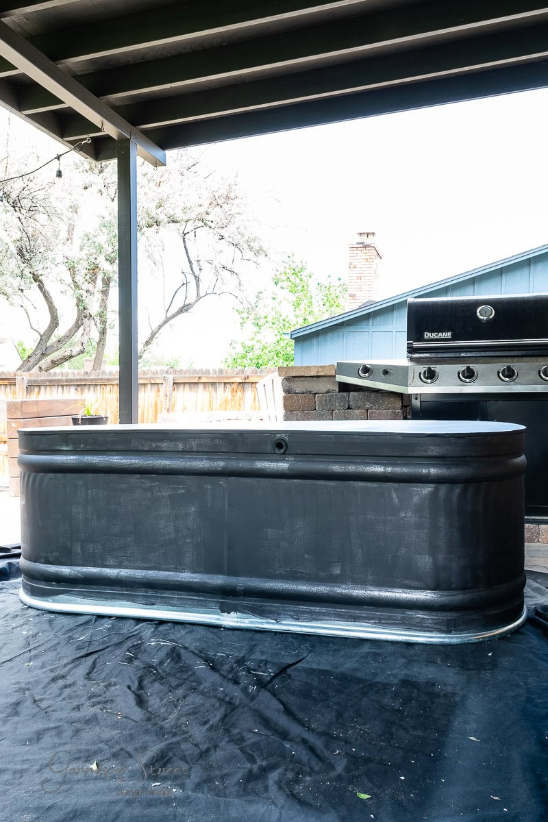 A large black metal tank, reminiscent of a stock tank, is placed under a covered patio with a barbecue grill nearby. This serene garden area is outdoors, surrounded by trees and a wooden fence. The patio roof is supported by sturdy metal beams.