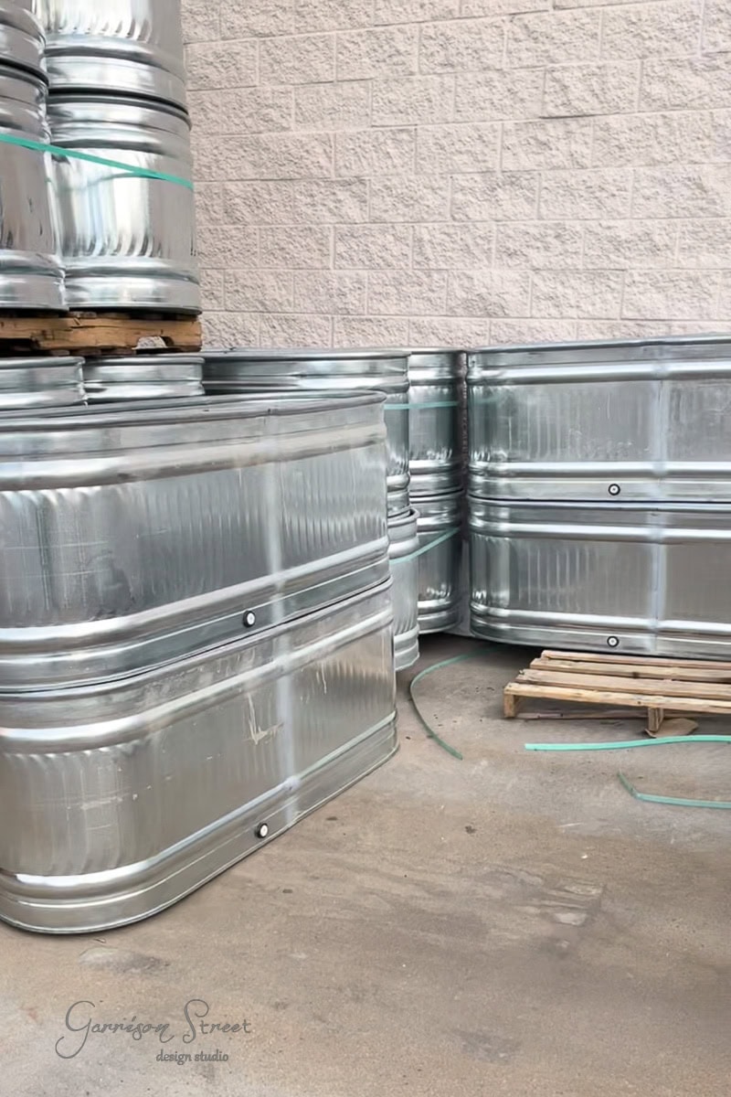 Several large, shiny silver stock tanks are stacked and placed on a concrete floor. A wooden pallet and a beige brick wall are visible in the background, evoking an industrial garden vibe.