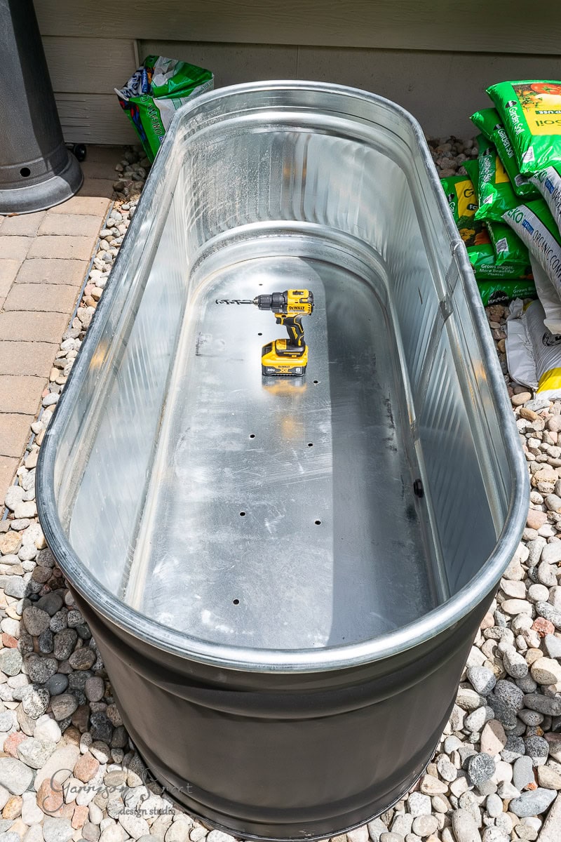 A large metal stock tank is placed on a bed of small rocks. Inside the tank, a yellow and black power drill is resting, ready for use. Bags of gardening soil are stacked in the background, creating a perfect garden setup.