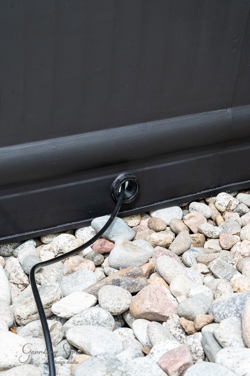 A black cable passes through a grommet at the base of a dark panel, surrounded by small garden stones.