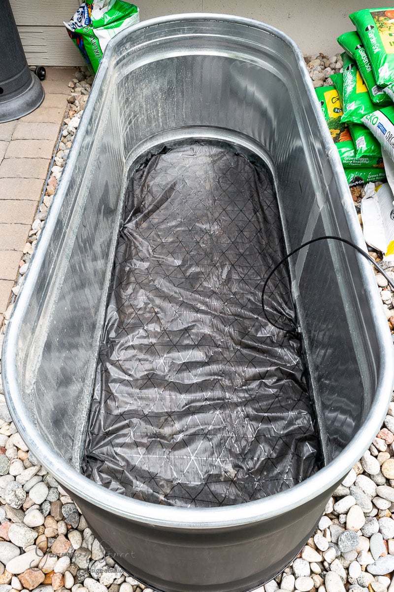 A large, empty stock tank with a black liner inside is placed on a bed of small stones, ready to be transformed into an exquisite garden feature. Bags of gardening soil are partially visible in the background, hinting at upcoming planting adventures.