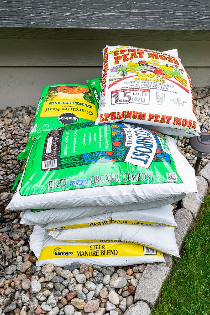 Bags of gardening materials, including peat moss, garden soil, organic compost, and steer manure blend, are stacked on a bed of rocks next to a thriving garden. Nearby, an old stock tank adds rustic charm to the picturesque scene.