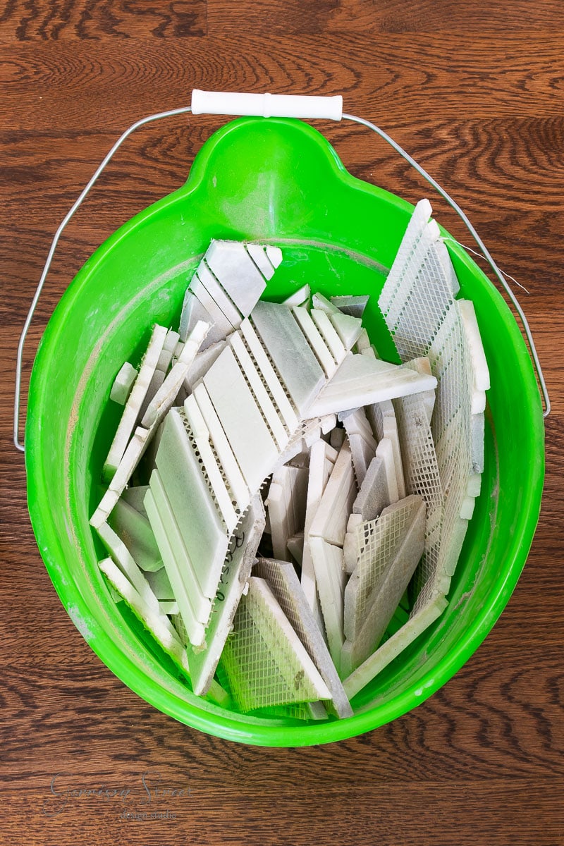 A green plastic bucket filled with broken white ceramic tile pieces, perfect for creating unique garden markers, rests on a wooden floor. The bucket features a wire handle with a white grip. The tiles are arranged in a random pattern within the bucket.