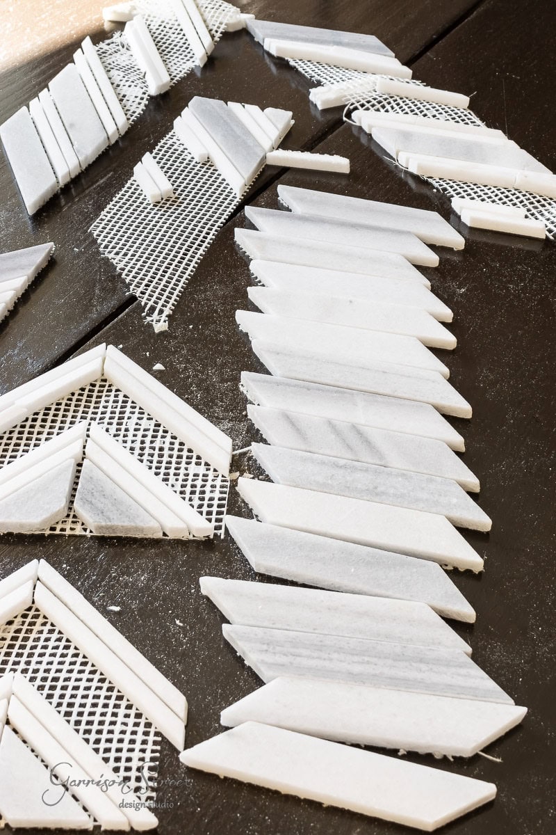 White marble tiles arranged in a herringbone pattern on a dark surface evoke an elegant style. Some are attached to mesh backing, while others are loose, resembling giant garden markers with their angular shapes and smooth texture.