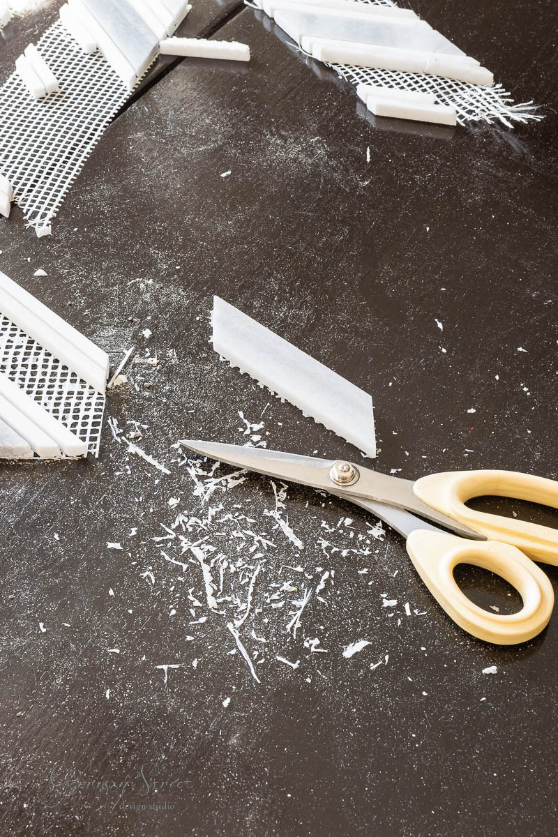 A pair of scissors rests on a dark surface surrounded by white plastic shavings and mesh pieces. Various strips of white plastic, perhaps being fashioned into garden markers, are scattered around, indicating a crafting or DIY project in progress.