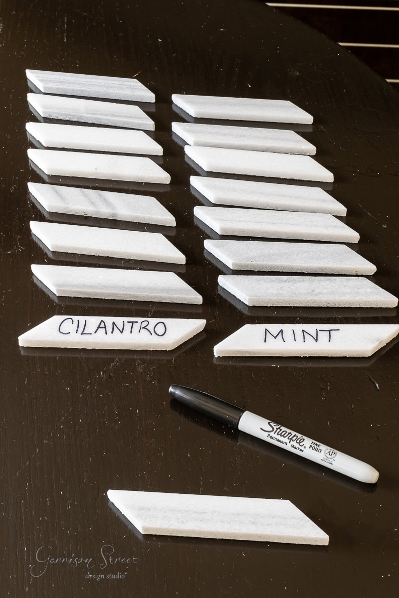 White garden markers with handwritten labels "CILANTRO" and "MINT" are arranged on a dark surface. A black Sharpie marker is nearby.