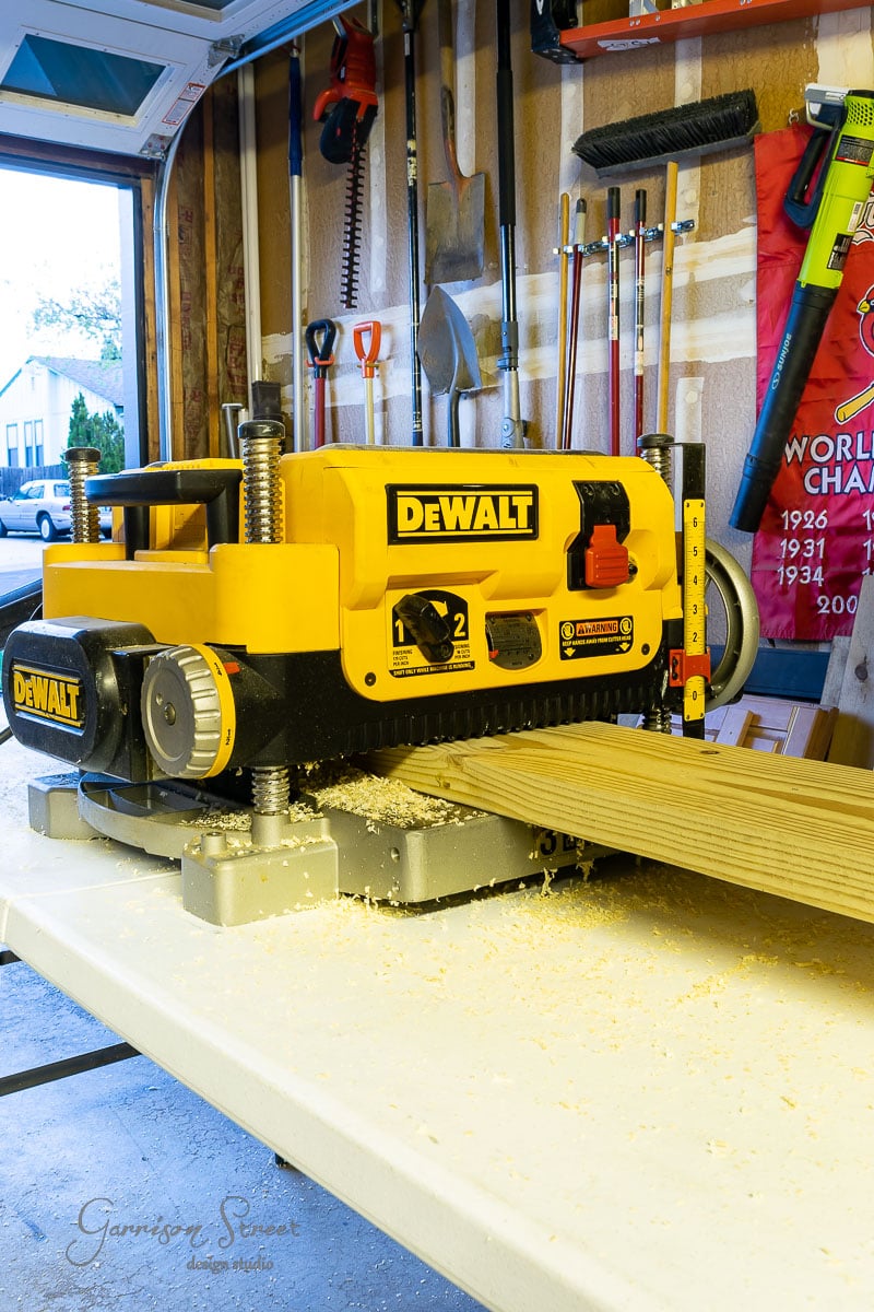 In a garage workshop, a DeWalt planer hums as it smooths a wooden board for a DIY wine rack project. Sawdust dances around the machine while tools hang neatly on a pegboard in the background, ready to bring your creation to life.