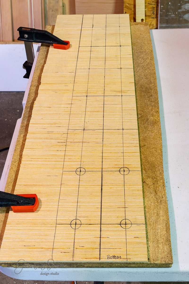 A wooden board clamped to a table with a grid drawn on its surface showcases the start of a DIY wine rack project. Circles at intervals indicate measured points for precision placement. Parts of the board and table are visible, with clamps ensuring stability during the build.