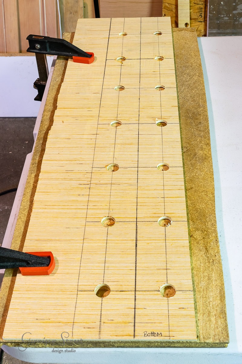 A wooden board clamped to a workbench, complete with drilled holes and penciled grid lines, sets the stage for learning how to build a DIY wine rack. Labels and a design studio logo add a professional touch to this intriguing crafting or woodworking setup.