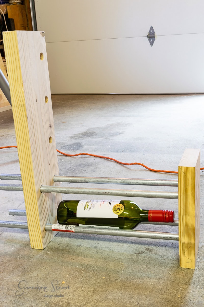 A wooden DIY wine rack in progress, held together with metal rods, rests on a concrete floor. A wine bottle sits securely on the bottom rods, while an orange extension cord snakes through the background, hinting at ongoing craftsmanship and creativity.