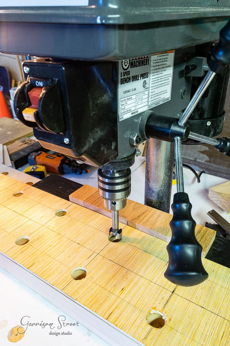 A bench drill press is shown in a workshop setting, drilling into a wooden board with evenly spaced holes—perfect for learning how to build a DIY wine rack. The machine is black with a metal spindle and chuck, set on a surface surrounded by various tools and hardware items.