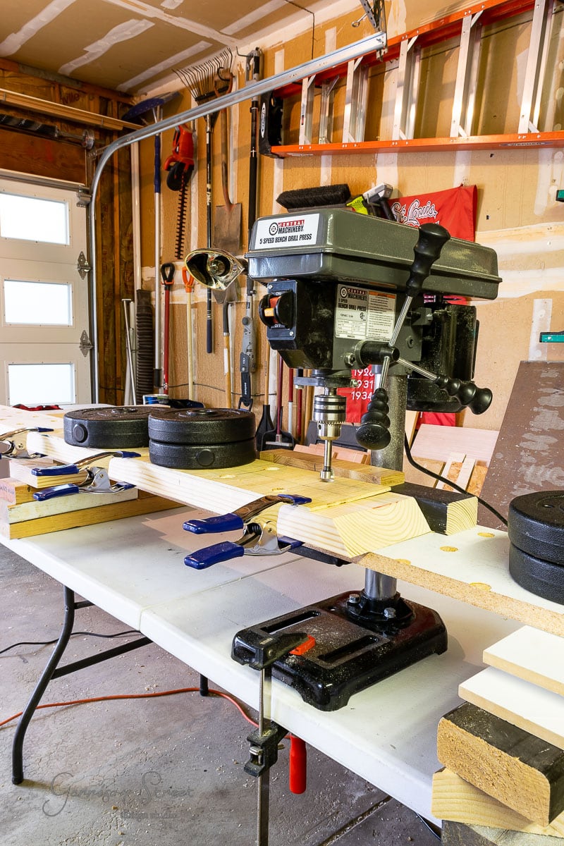 A garage workshop serves as the creative hub for building projects like a DIY wine rack. A drill press sits on a table cluttered with wooden boards, clamps, and weights. Tools are scattered in the background alongside a ladder and the white garage door, ready for the next crafting session.