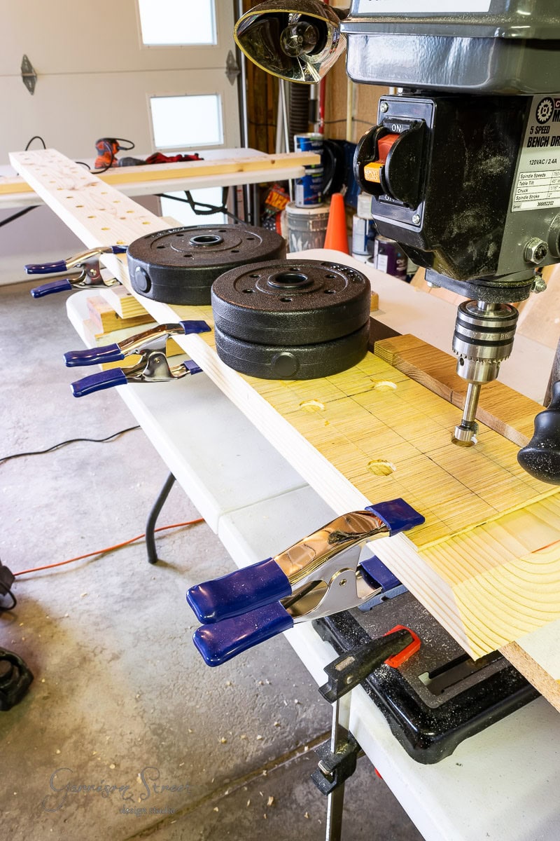 A wooden board is secured on a table with blue clamps, surrounded by weight plates for stability in a busy workshop. A drill press hovers above, ready to assist in your next project: how to build a DIY wine rack with precision and style amidst the array of tools and equipment. .