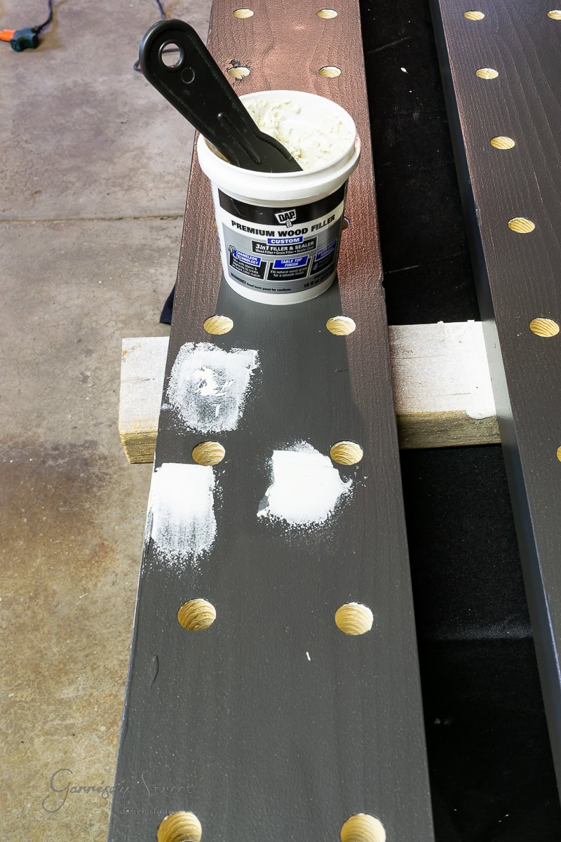 A wooden beam with evenly spaced circular cutouts sits on supports, resembling the start of a DIY wine rack. Partly painted gray with white wood filler applied, it has a putty knife resting on an open container of wood filler atop the beam.