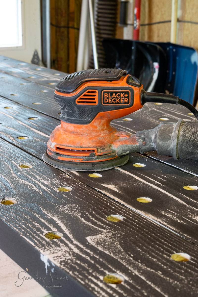 In a workshop setting, an orange and black Black+Decker orbital sander suavely smooths the textured tabletop, shaping it for a DIY wine rack project. Its circular indents are ideal for ensuring a quality finish on your handmade creation.