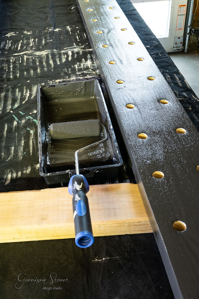 A paint roller rests in a tray of black paint on a covered surface, poised for action. Nearby, a long wooden board with evenly spaced holes hints at an intriguing project—perhaps the creation of a chic wine rack. The scene unfolds like the beginning steps in building your own DIY masterpiece.
