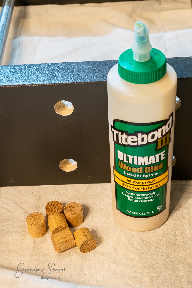 A bottle of Titebond Ultimate Wood Glue stands ready for action beside small wooden dowels and a piece of dark wood with circular holes, set on a light-colored fabric—a perfect setup for those looking into how to build a DIY Wine Rack.