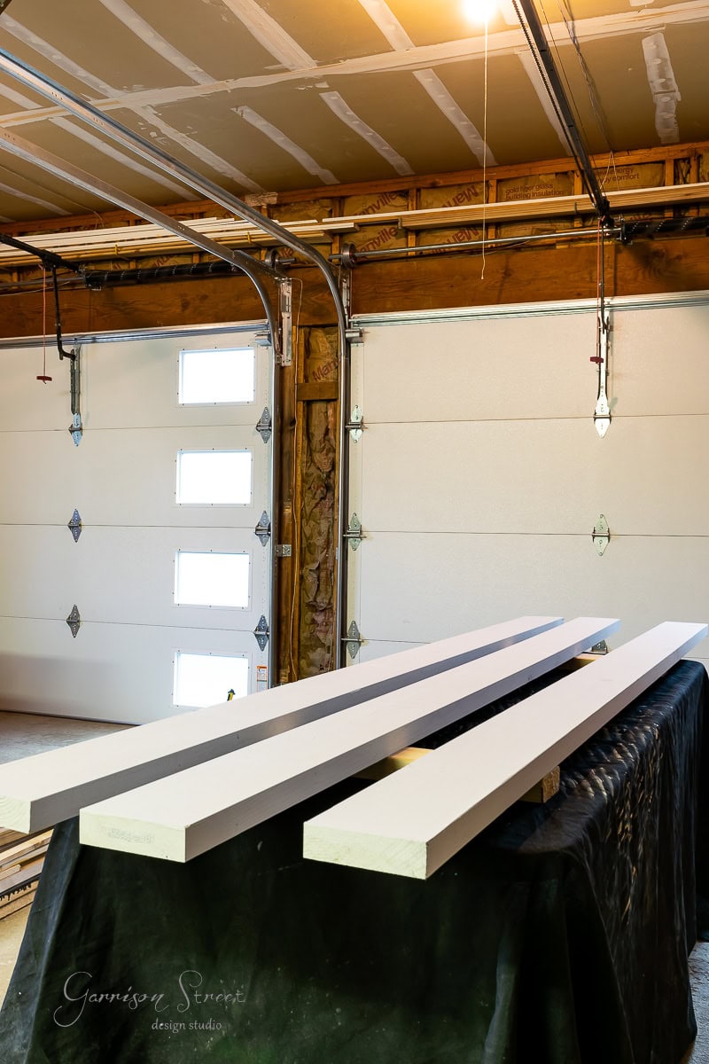 Three long white wooden boards rest on a black covered table in a garage, setting the stage for a "How to build a DIY Wine Rack" project. The backdrop features a closed garage door with small windows, unfinished walls, and ceiling lights—perfect for crafting your masterpiece.