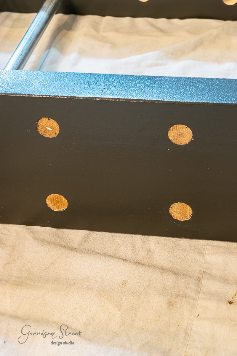 Close-up of a black metal piece with four round holes arranged in a square pattern—perfect for your next "How to Build a DIY Wine Rack" project. Placed on beige fabric, the logo "Garrison Street Design Studio" graces the bottom left corner, adding an elegant touch.