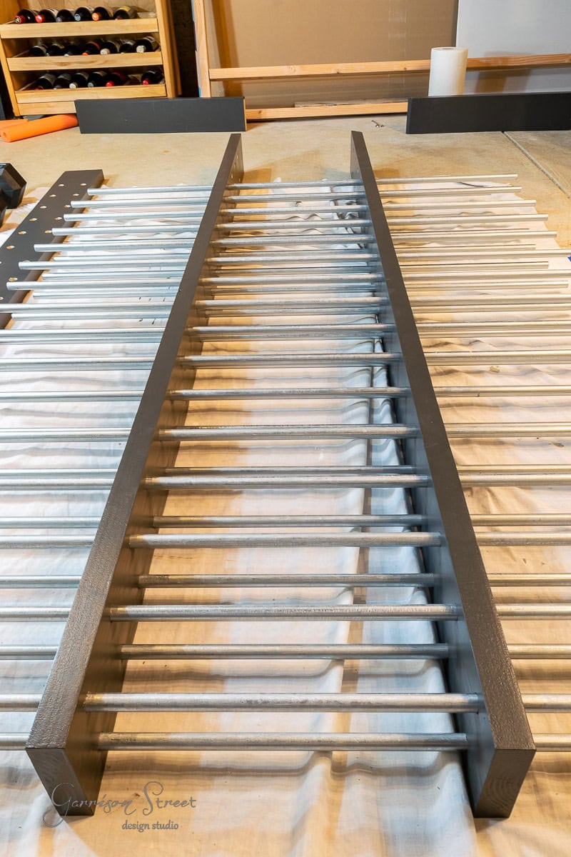 A wooden baby gate painted in gray and white with vertical slats is laid flat on a covered floor, surrounded by various painting supplies and tools, reminiscent of a workshop where one might learn how to build a DIY wine rack.