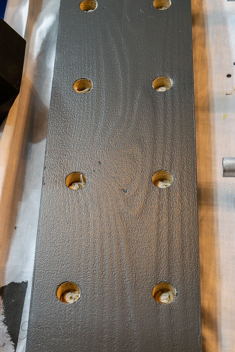 A close-up of a textured gray panel with eight evenly spaced circular holes, ideal for learning how to build a DIY wine rack. Wood shavings are visible within each hole, indicating recent drilling. The panel rests on a light-colored surface, ready for assembly.