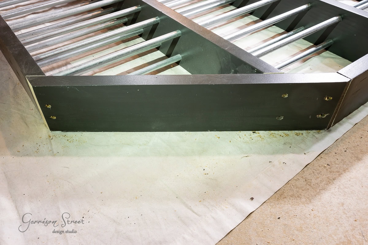 A metal bed frame rests on a light-colored carpet, its dark finish complemented by sturdy metal slats. A glimpse of a drop cloth or sheet peeks from underneath. In the corner, an obscured logo adds intrigue, much like figuring out how to build a DIY wine rack at home.