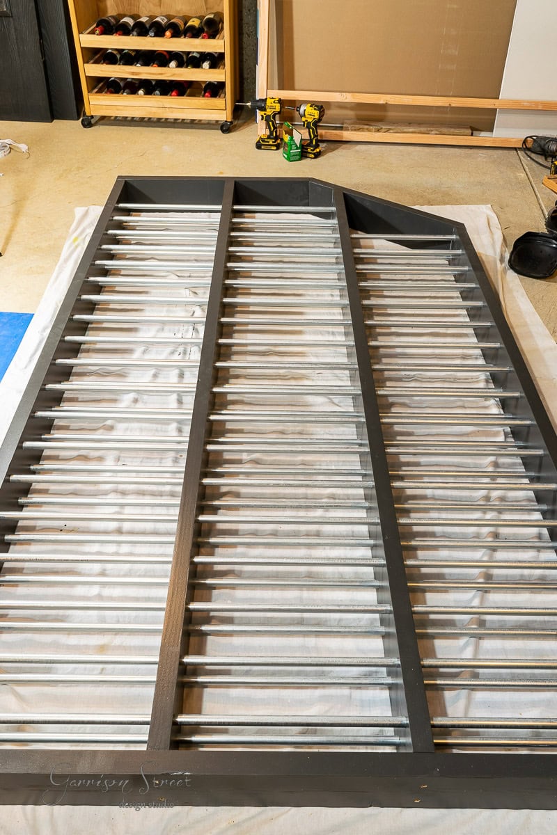 A large gray metal gate or railing lays flat on a white sheet in a workshop, amidst an array of tools essential for DIY projects. Nearby, a shelf with paint bottles, a cordless drill, and a wooden board—perfect for crafting your next project like building a stunning DIY wine rack—lean against the wall.